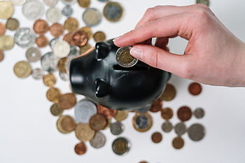 Close-up of a hand inserting a coin into a black piggy bank with scattered coins on a white background. HD wallpaper