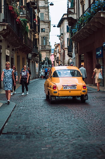 Charming scene of a vintage yellow car driving down a cobblestone street in Ferrazze, Italy, with pedestrians walking nearby. HD phone wallpaper