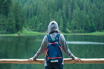 A person in a hoodie stands by a serene lake surrounded by lush greenery, enjoying the peaceful outdoors. HD wallpaper