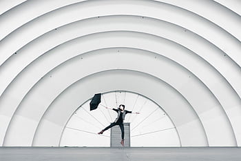 Person jumping joyfully under large architectural arches with an umbrella. HD wallpaper