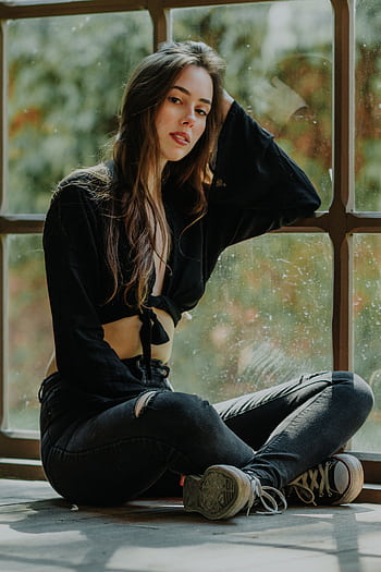 Fashionable young woman sitting by a window in casual attire, exuding confidence and style. HD phone wallpaper