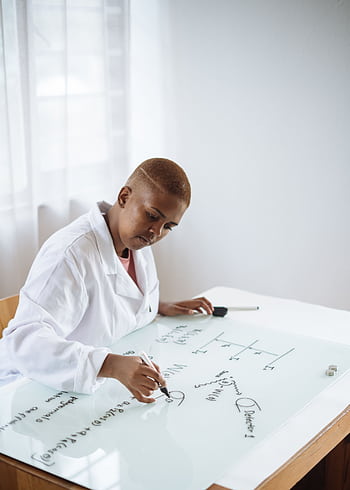 African-American female scientist in lab coat writing scientific equations on a glass board. HD phone wallpaper