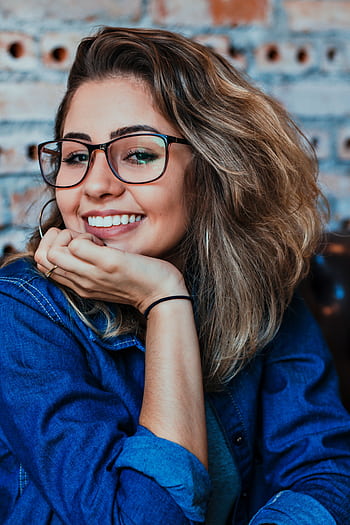 A young woman wearing glasses and denim, smiling in an indoor setting. HD phone wallpaper