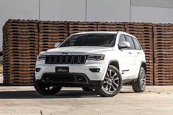 A sleek white Jeep Grand Cherokee parked in front of stacked wooden pallets, showcasing its luxury features. HD wallpaper