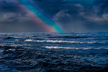 Mesmerizing ocean waves with a vibrant rainbow and surfers under a cloudy sky. HD wallpaper