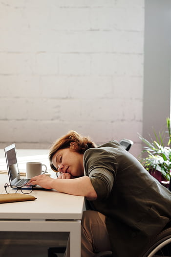 A tired woman in a wheelchair sleeps at her desk in a modern office setting. HD phone wallpaper