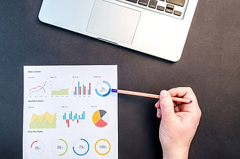 Hand holding pencil reviewing colorful data charts on desk with laptop. HD wallpaper