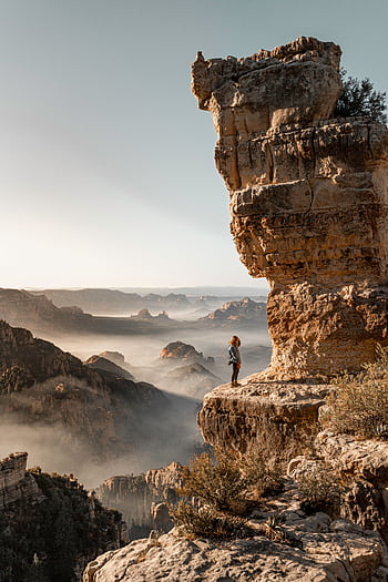 A lone traveler stands on a rocky cliff, overlooking a breathtaking mountain landscape filled with mist and morning light. HD phone wallpaper