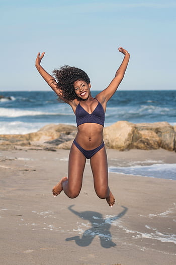 A woman in swimwear joyfully jumps on a sunny beach, exuding happiness. HD phone wallpaper