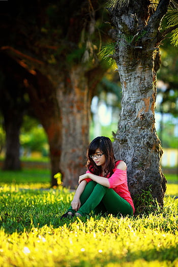 A young woman in vibrant attire sits pensively under a tree in a sunlit park. HD phone wallpaper