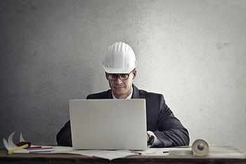 Professional engineer wearing hardhat working on construction plans at desk with laptop in office. HD wallpaper