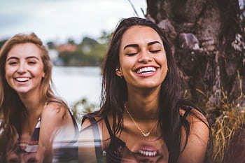 Two women happily laughing together outdoors by a serene lakeside. HD wallpaper