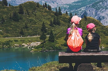 Two young girls sitting on a bench overlooking a serene mountain lake, enjoying a summer hike. HD wallpaper