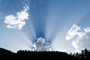 A stunning natural phenomenon as sunrays pierce through fluffy clouds above a silhouetted forest. HD wallpaper