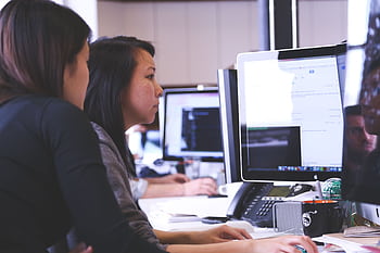 Two women working together on code at a computer in a modern office setting. HD wallpaper