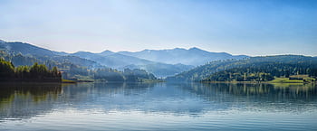 A tranquil mountain lake surrounded by mist-covered forests and distant peaks under a clear blue sky. HD wallpaper
