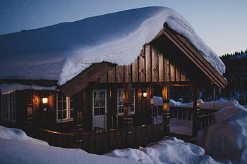 A snow-covered wood cabin illuminated warmly against a winter dusk sky, surrounded by snowy landscape. HD wallpaper