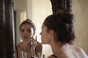 A woman checks her skin in the mirror, engaging in her morning skincare routine indoors. HD wallpaper