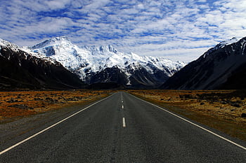 A deserted road leading to majestic snow-covered mountains under a cloudy blue sky. HD wallpaper