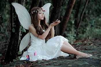 A captivating of a fairy in white dress with wings, sitting in a woodland setting, holding a butterfly. HD wallpaper