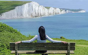 A woman relaxes on a bench, admiring the scenic cliffs and ocean view. HD wallpaper