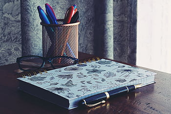 A stylish workspace featuring a floral notebook, glasses, and pens on a wooden desk. HD wallpaper