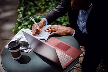 Man writing notes in a notebook while working on a laptop outdoors, symbolizing remote work lifestyle. HD wallpaper