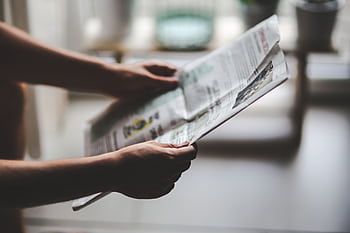 Hands holding a newspaper in a soft-lit, indoor environment, focusing on a relaxed reading moment. HD wallpaper