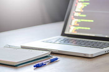 Open laptop with programming code on screen next to a notebook and pen on a desk. HD wallpaper