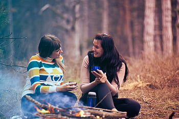 Two women enjoy coffee by a campfire in a forest, embracing the warmth and togetherness of nature. HD wallpaper