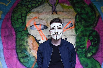 A person in a Guy Fawkes mask stands before colorful graffiti art, creating an air of mystery. HD wallpaper