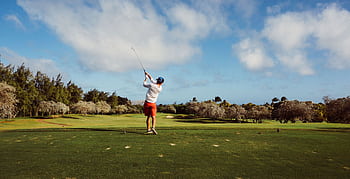 A golfer swings on a que golf course with lush greenery and a clear blue sky, epitomizing outdoor leisure. HD wallpaper
