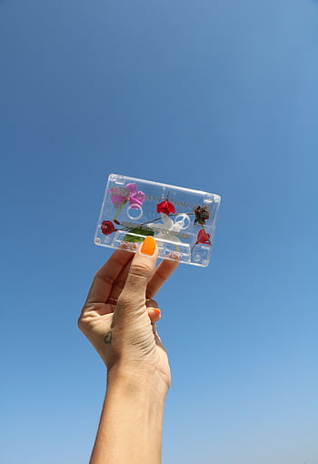 A hand with painted nails holds a flower-decorated cassette against a bright clear blue sky, symbolizing nostalgia and creativity. HD phone wallpaper