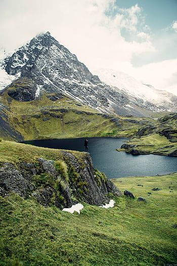 A hiker stands on a cliff overlooking a serene mountain lake surrounded by lush greenery and snow-capped peaks. HD phone wallpaper