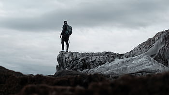 A lone hiker stands at the edge of a rugged cliff with a moody sky overhead, epitomizing adventure. HD wallpaper