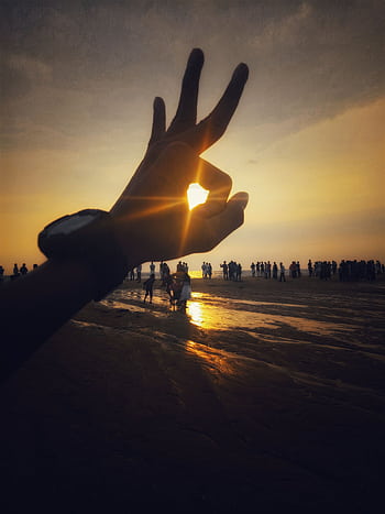 A hand gesture silhouetted against a vibrant sunset on a crowded beach. HD phone wallpaper