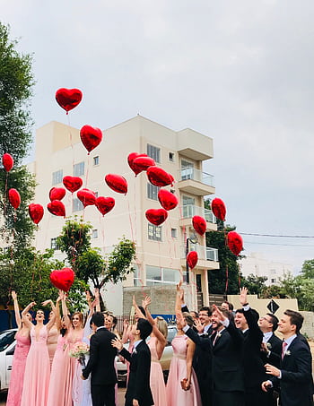 A joyful wedding party celebrating outdoors with red heart balloons in the air against an urban backdrop. HD phone wallpaper