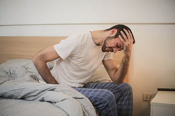 A young man in pajamas holding his head, sitting on a bed, appears to be experiencing a headache. HD wallpaper