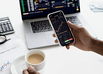 A person trades stocks on a smart with a laptop, coffee cup, calculator, and documents in view. HD wallpaper