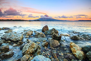 A scenic view of a sunset over a volcanic island, with rocks in the foreground and a calm sea. HD wallpaper
