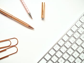 Elegant minimalist workspace featuring rose gold pens, paper clips, and a sleek keyboard for a stylish office setup. HD wallpaper