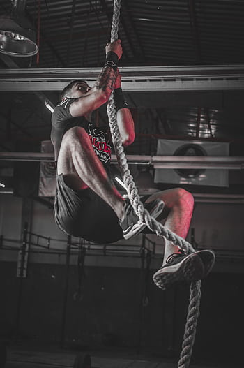 A determined man climbing a rope in an indoor gym setting, showcasing strength and fitness. HD phone wallpaper