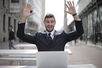 A joyful businessman raises his hands in excitement while working on a laptop outdoors. HD wallpaper