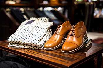 Elegant brown leather shoes and printed shirt displayed on a wooden table in a stylish clothing store. HD wallpaper