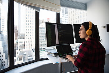 A businesswoman coding at a standing desk with city views through large windows, wearing heads. HD wallpaper