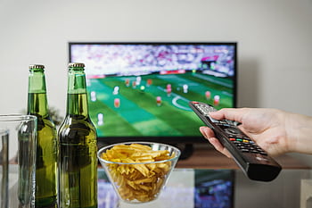 A person holding a remote while watching soccer on TV with beer and snacks on the table. HD wallpaper