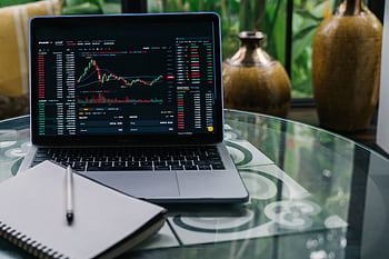 Laptop displaying cryptocurrency stocks and graphs on a glass table with a notepad. HD wallpaper
