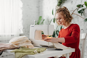 A woman in a red dress using a sewing machine at home, creating a handmade garment. HD wallpaper