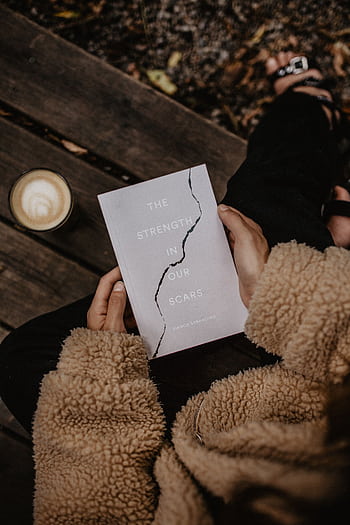 Woman reading 'The Strength in Our Scars' book outdoors in autumn with coffee. HD phone wallpaper