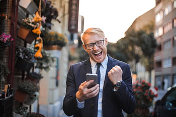 A joyful man in a suit celebrates good news on his smart in an urban street setting. HD wallpaper
